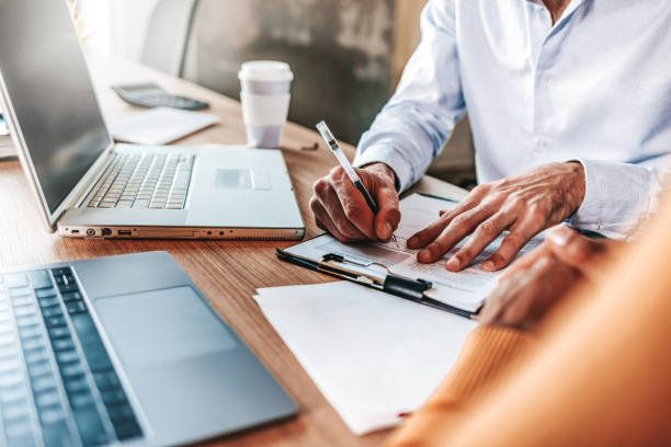 Couple reading legal documents at home with laptop, family considering mortgage loan or insurance, studying contract details, discussing terms and conditions, close up view of hands holding papers.