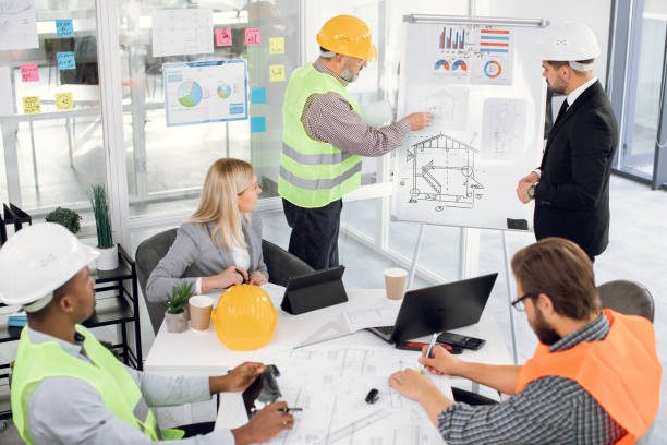 Focused bearded male chief in suit listening to professional middle aged bearded man builder contractor about building plan on blueprint from during meeting in boardroom