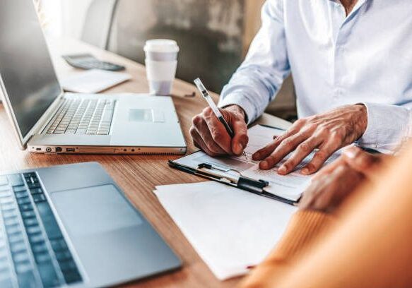 Couple reading legal documents at home with laptop, family considering mortgage loan or insurance, studying contract details, discussing terms and conditions, close up view of hands holding papers.